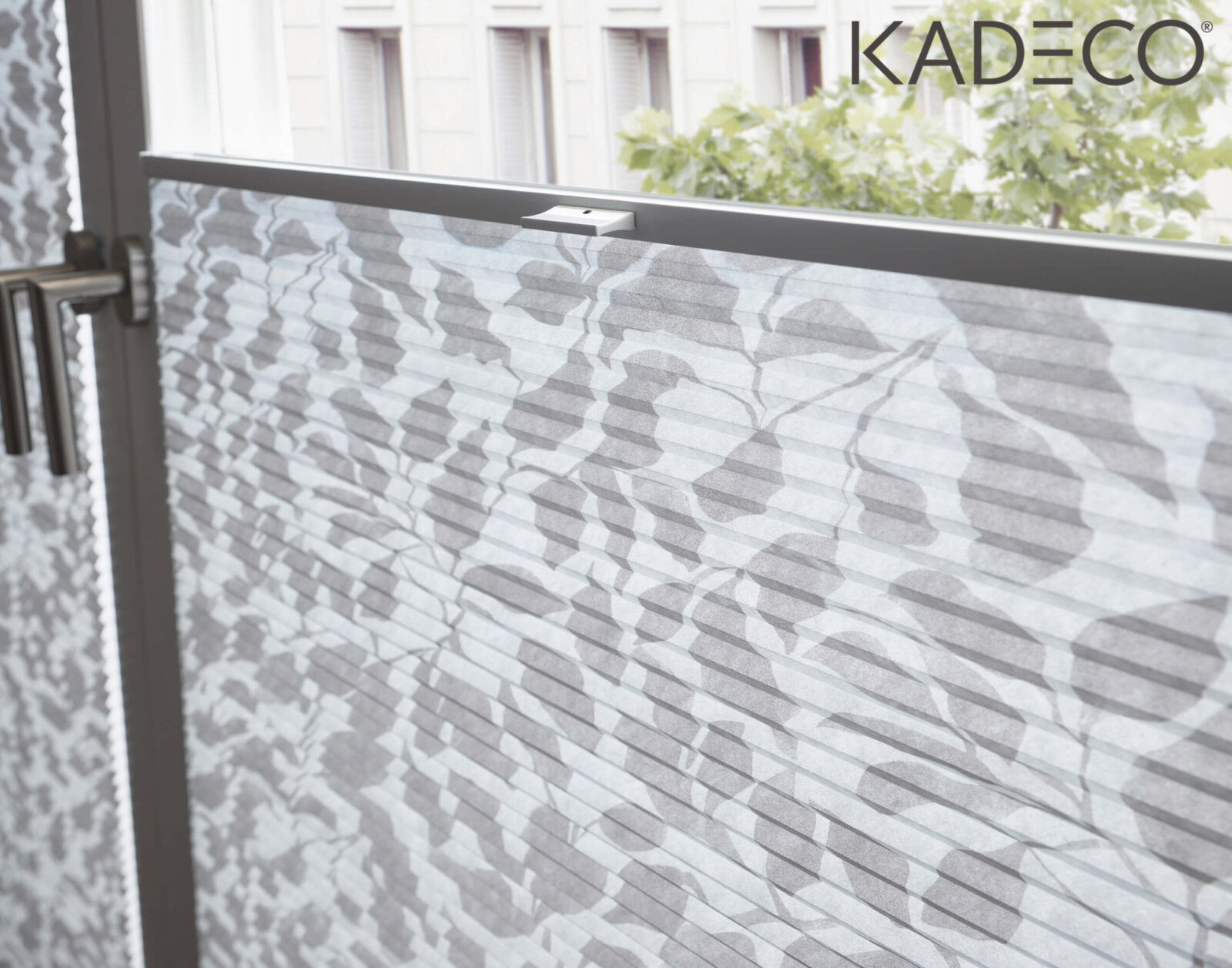 Ein graues Wabenplissee von KADECO mit filigranem Blattmuster dient als optisches Highlight